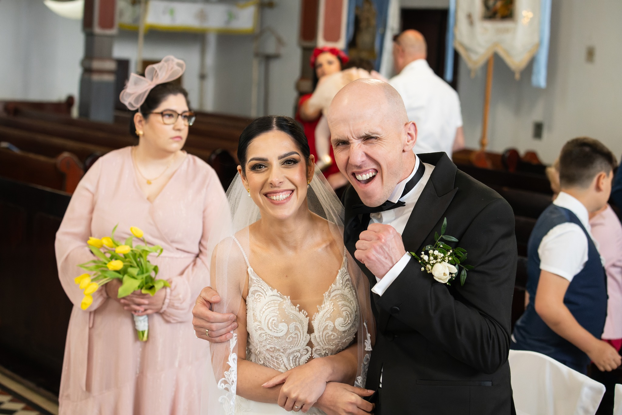 Uśmiechnięta panna młoda w koronkowej sukni i pan młody w smokingu z zabawną miną, obok druhna z żółtymi tulipanami w kościele.