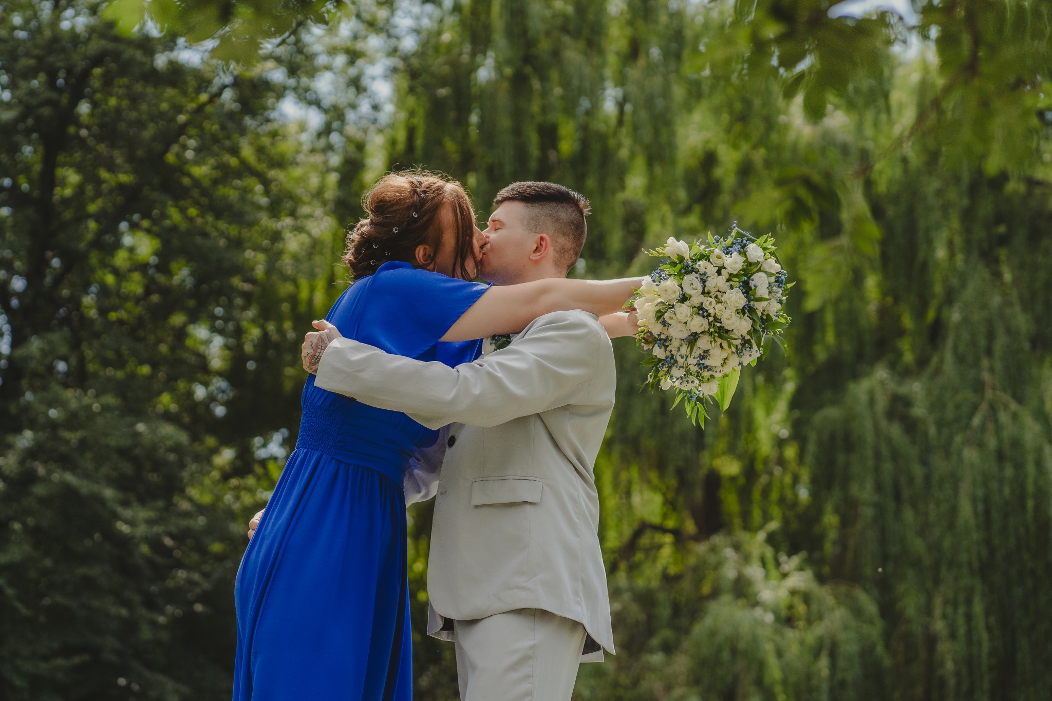 Para młoda w objęciach, panna młoda w kobaltowej sukience, pan młody w jasnym garniturze, bukiet z białych róż i niebieskich akcentów, pocałunek na tle zieleni drzew.