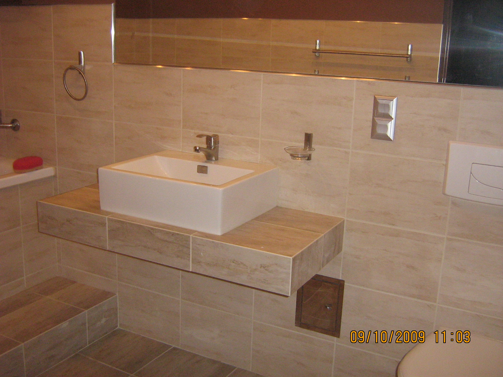 Łazienka po remoncie: kwadratowa umywalka nablatowa na blacie z płytek imitujących marmur, beżowe płytki na ścianach i podłodze, widoczny fragment toalety.