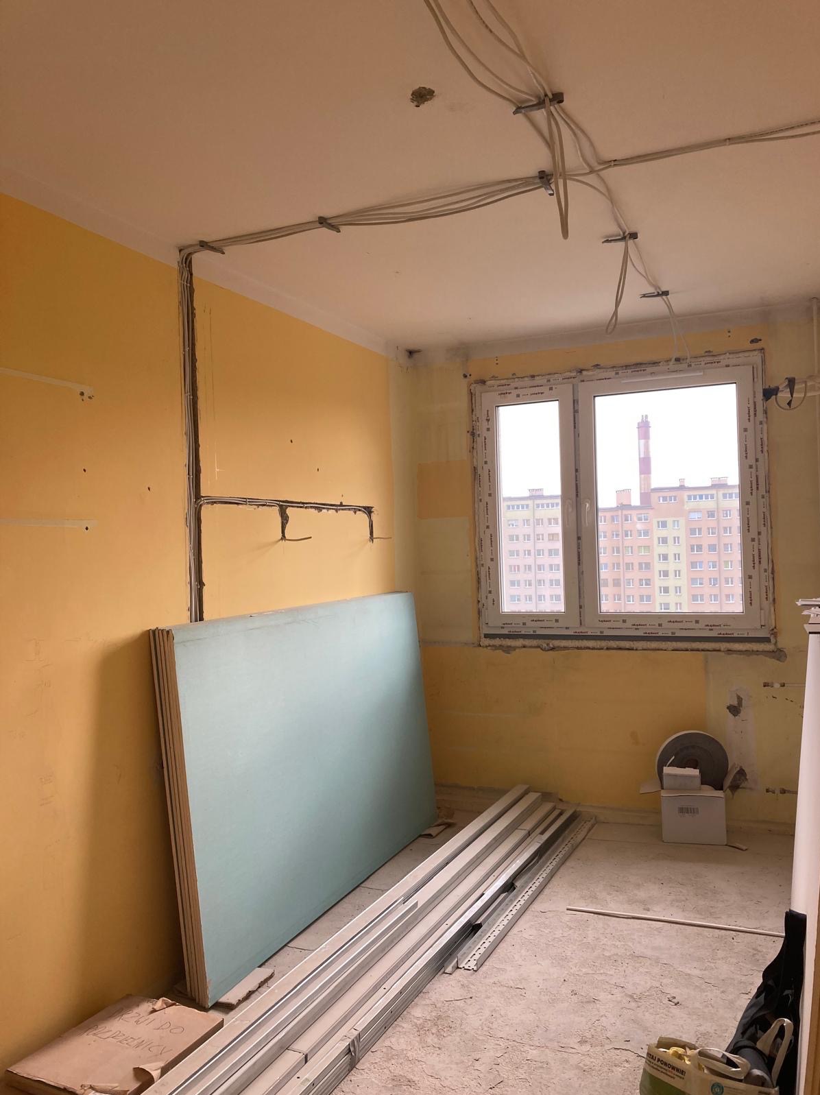 Pokój w trakcie remontu: żółte ściany, płyty gipsowe oparte o ścianę, profile metalowe na podłodze, widoczne kable elektryczne pod sufitem. Okno z widokiem na blok.