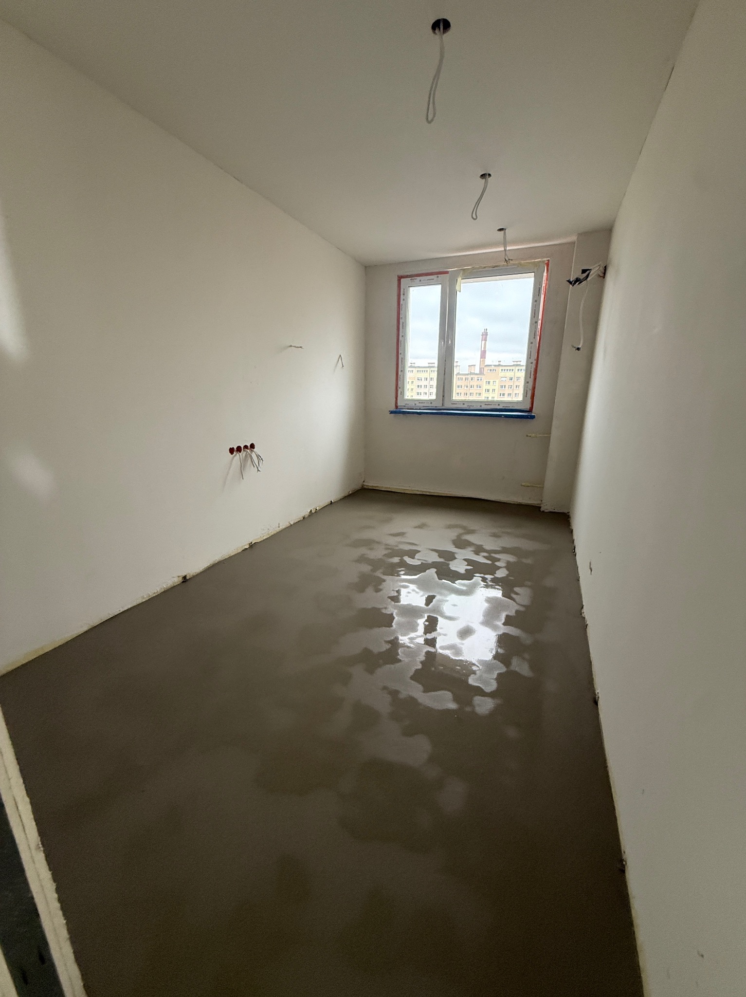 Wnętrze pokoju w stanie surowym z wylewką podłogową, widoczne instalacje elektryczne i okno z widokiem na miasto. Świeżo wylana, mokra podłoga.