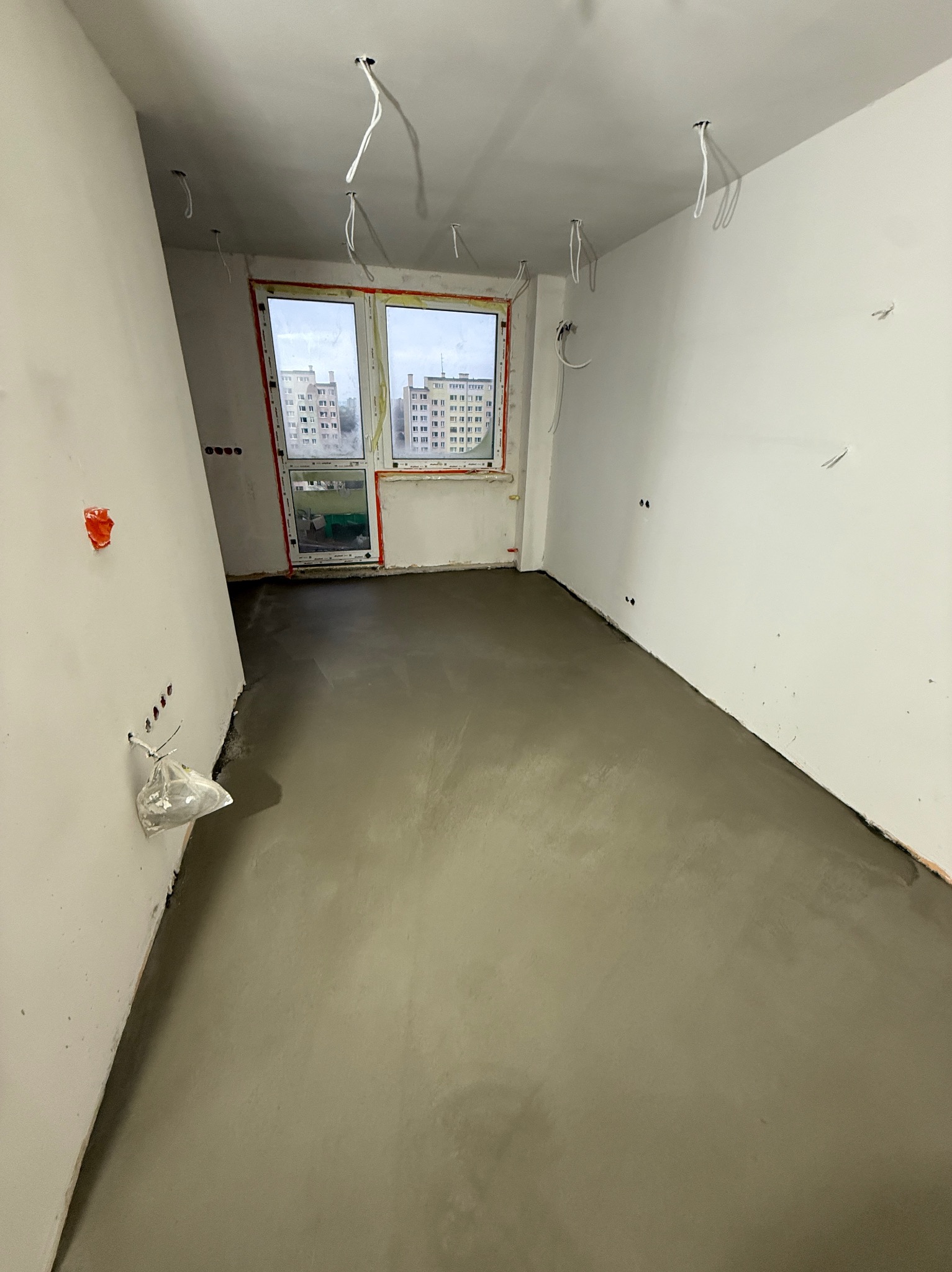Wnętrze po wylaniu posadzki: świeża, szara wylewka na podłodze, białe ściany z wystającymi kablami, okno z widokiem na blok. Etap prac wykończeniowych.