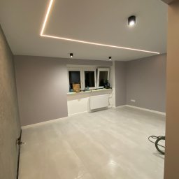 Yonex - Pomalowane na liliowo ściany, jasna podłoga z płytek, okno z białym parapetem, grzejnik, czarne lampy sufitowe i kwadratowe oświetlenie LED na suficie w pustym pokoju.