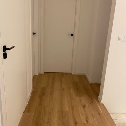 Yonex - Korytarz z nowo ułożonymi panelami podłogowymi w odcieniach jasnego dębu, widoczne białe drzwi z czarnymi klamkami i fragment ściany z gniazdkiem elektrycznym.