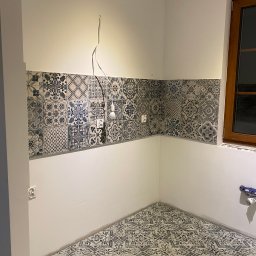 Yonex - Kuchnia w trakcie remontu: ułożone płytki dekoracyjne na podłodze i nad blatem, widoczne kable i gniazdka elektryczne, drewniane okno z parapetem.