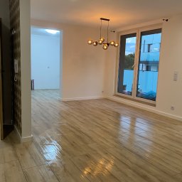 Yonex - Jasne, przestronne wnętrze mieszkania w Katowicach po remoncie, z drewnopodobną podłogą z paneli, nowoczesną lampą wiszącą i dużym oknem z widokiem na zewnątrz.