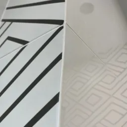 Styk dwóch rodzajów płytek: biała z szarym geometrycznym wzorem i biała z czarnymi ukośnymi pasami, widoczne delikatne niedoskonałości fugi.