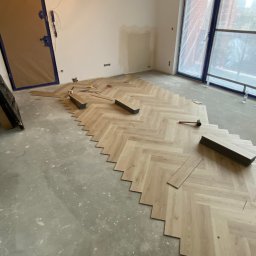 Yonex - Układanie jasnych paneli podłogowych w jodełkę francuską, widoczne narzędzia i niedokończona podłoga w surowym wnętrzu z zabezpieczonymi drzwiami i oknami.