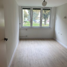 Yonex - Pusty pokój z nową podłogą z paneli, świeżo pomalowanymi ścianami na biało i trzema oknami z widokiem na zieleń na zewnątrz.