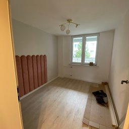 Yonex - Pusty pokój z panelami ściennymi w kolorze pudrowego różu, drewnianą podłogą i białymi ścianami, widok zza uchylonych drzwi, na podłodze leżą profile przypodłogowe i torba.