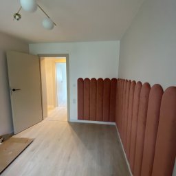 Yonex - Pusty pokój z jasną podłogą i dekoracyjnymi, pionowymi panelami ściennymi w odcieniach terakoty, zakończonymi zaokrąglonymi brzegami, widoczne niedokończone podłogi i otwarte drzwi.