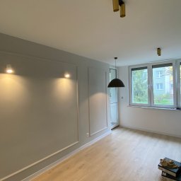 Yonex - Wnętrze pokoju po remoncie: szare ściany z dekoracyjnymi listwami, jasna drewniana podłoga, nowoczesne oświetlenie sufitowe, widok na zieleń za oknem, kartony na podłodze.