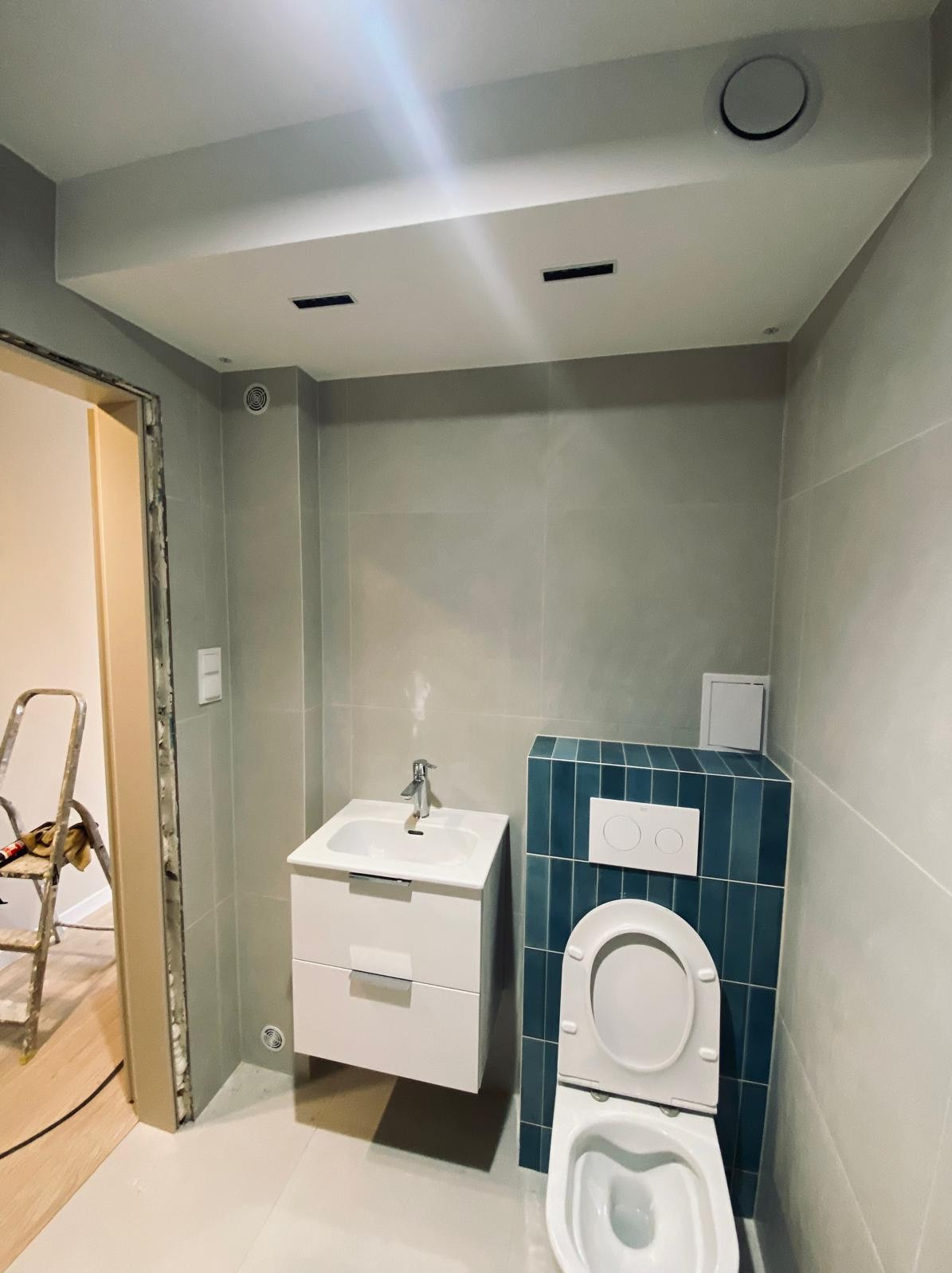 Nowoczesna łazienka w trakcie wykańczania: szare płytki ścienne, biała umywalka z szafką, toaleta obudowana niebieskimi płytkami, drabina malarska w tle przy niedokończonych drzwiach.