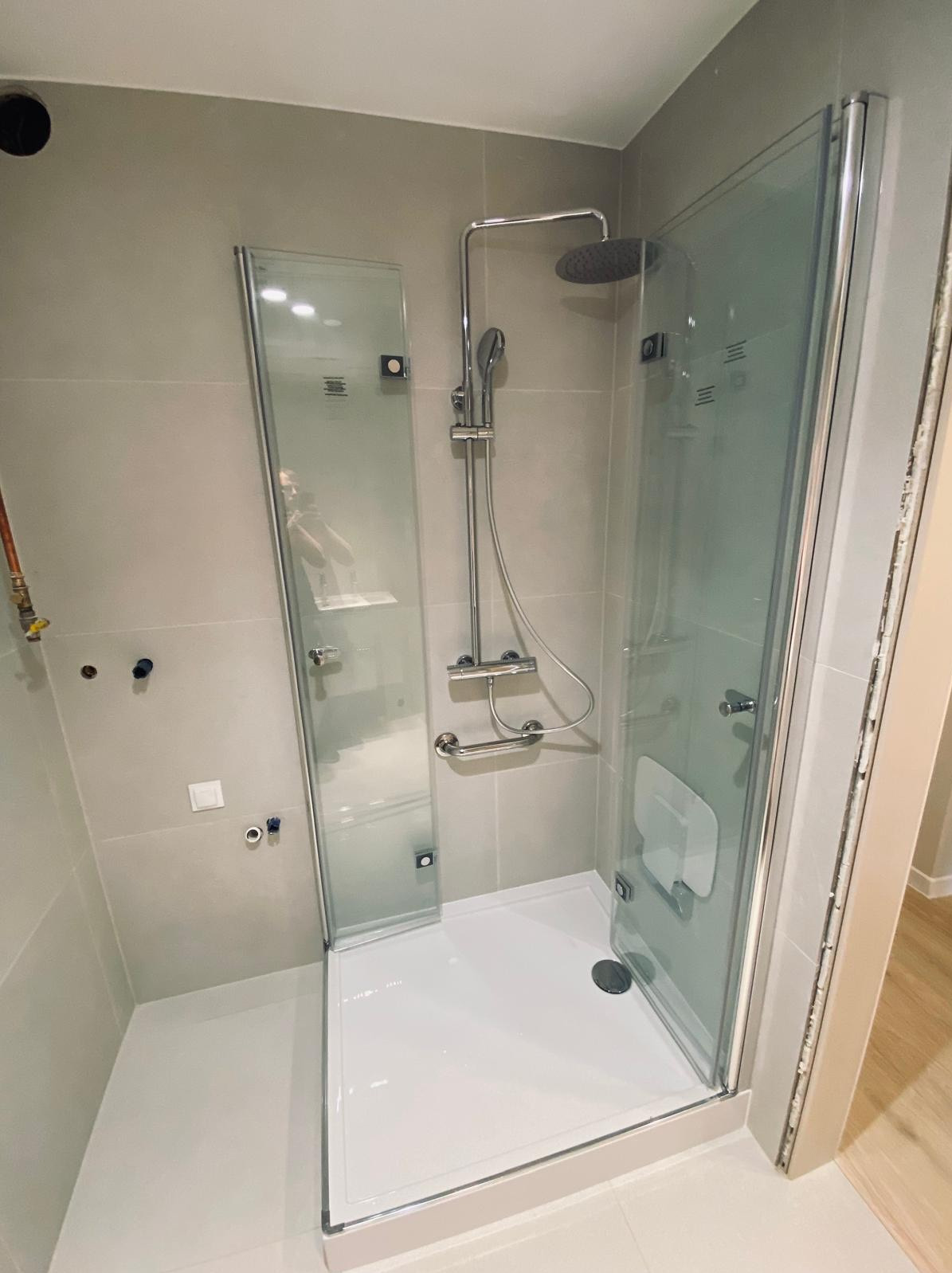 Półotwarta szklana kabina prysznicowa z siedziskiem i uchwytem dla niepełnosprawnych, widoczne niedokończone fugi przy drzwiach, szare płytki ścienne, instalacja wodna bez baterii.