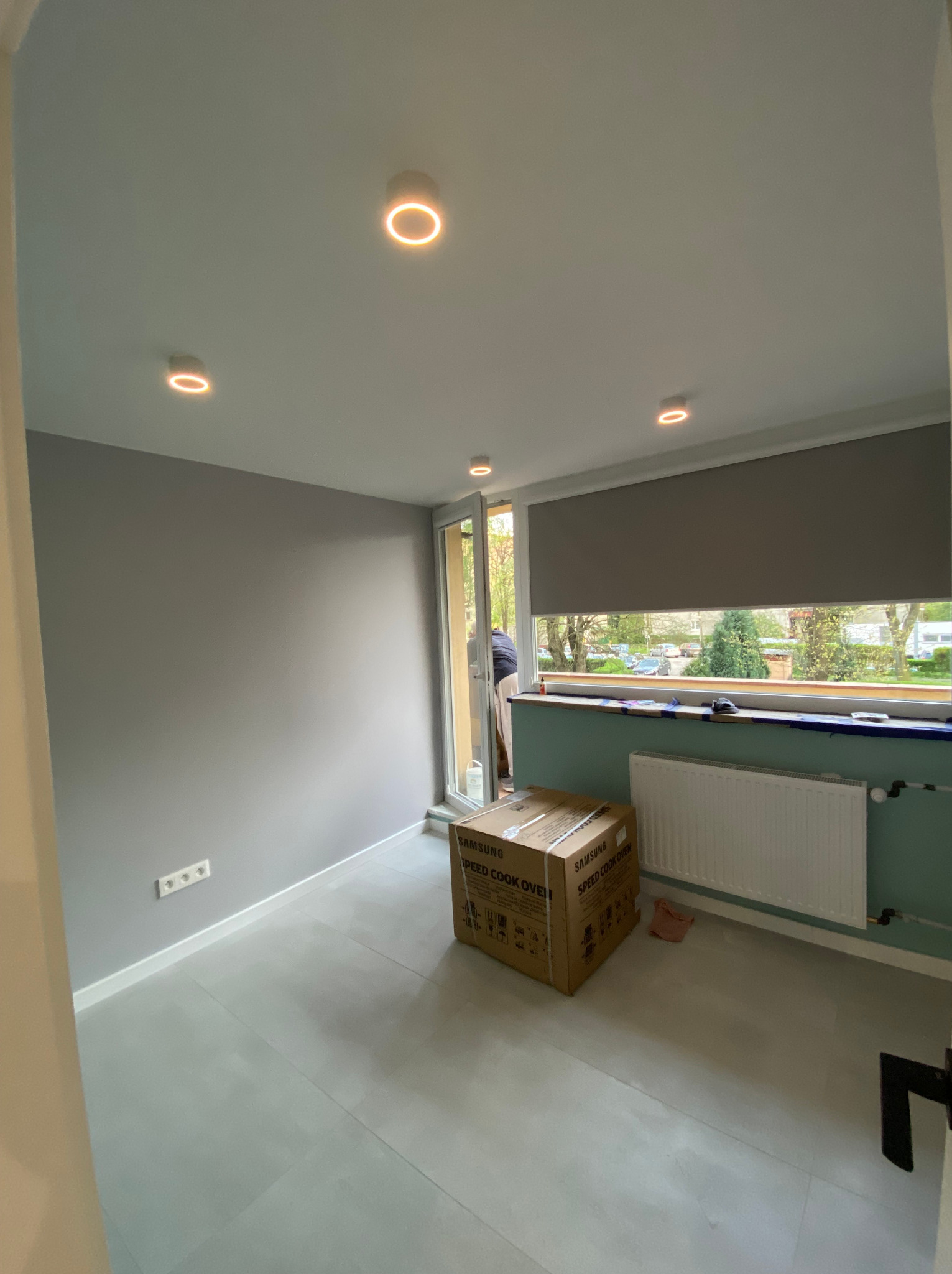 Szare wnętrze pokoju po remoncie z zamontowanymi okrągłymi lampami sufitowymi, grzejnikiem pod oknem i kartonem po urządzeniu AGD na podłodze