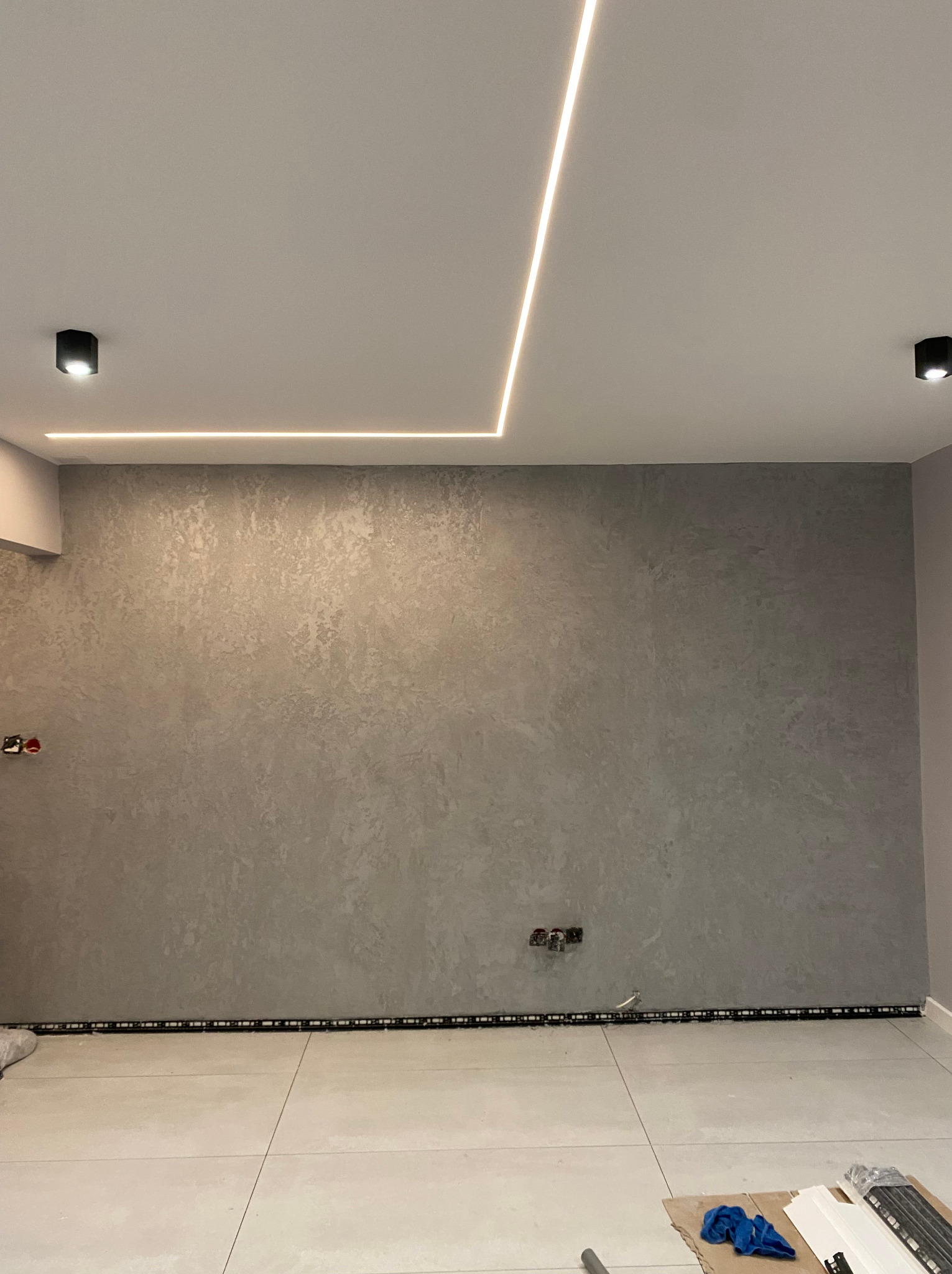 Ściana wykończona dekoracyjną szarą gładzią z widocznymi włącznikami i gniazdkami, podświetlona liniowym oświetleniem LED na suficie, z fragmentem podłogi z płytek i elementami wykończeniowymi...