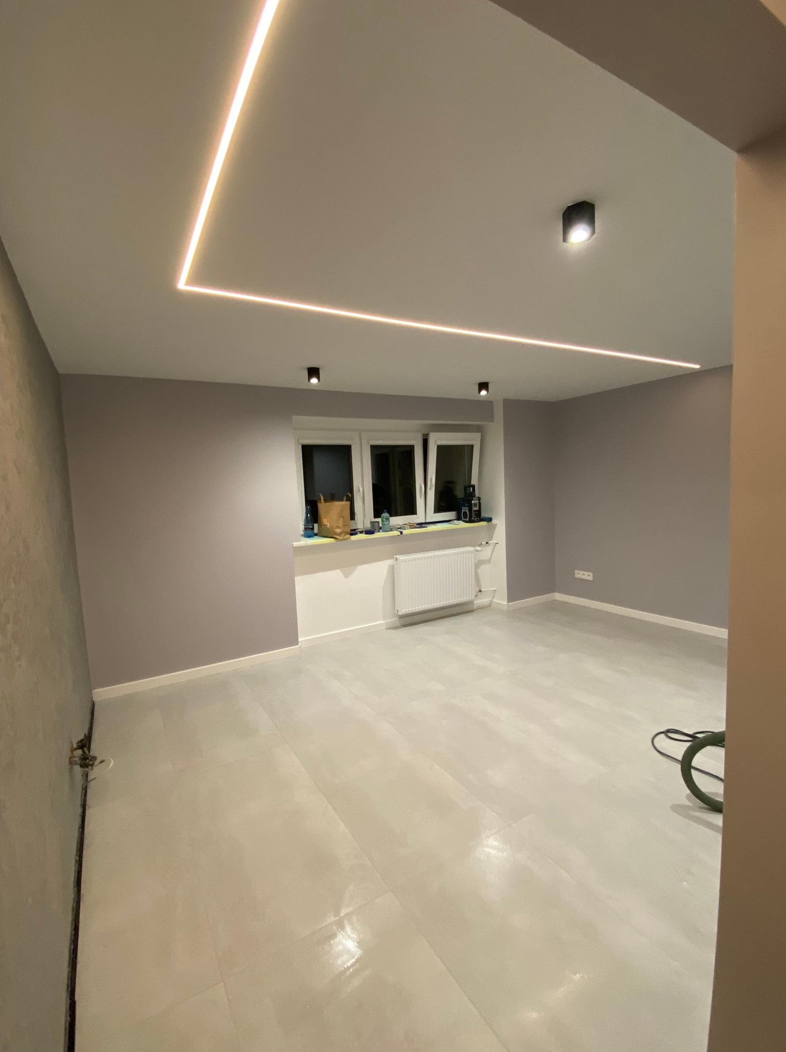 Pomalowane na liliowo ściany, jasna podłoga z płytek, okno z białym parapetem, grzejnik, czarne lampy sufitowe i kwadratowe oświetlenie LED na suficie w pustym pokoju.