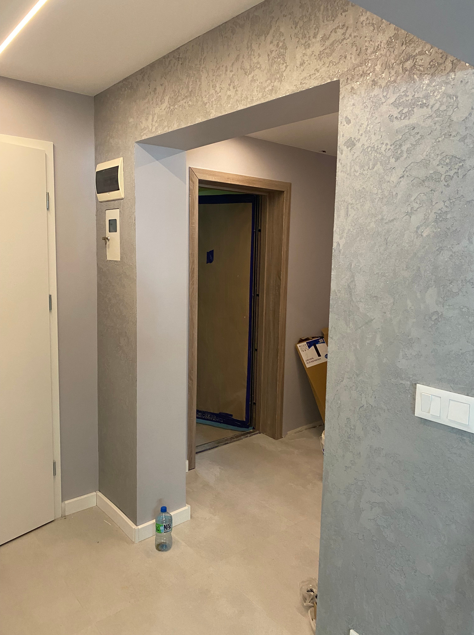 Korytarz po nałożeniu dekoracyjnej szarej gładzi z efektem strukturalnym na ścianach, widoczna butelka wody przy listwie przypodłogowej i zabezpieczone drzwi wejściowe.