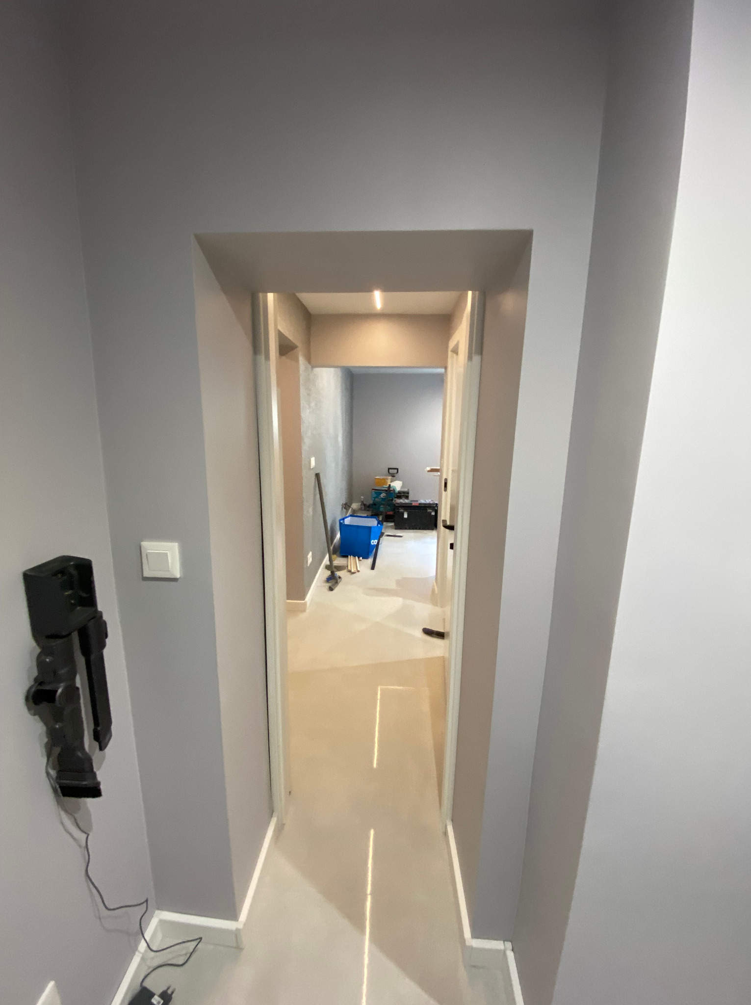 Korytarz w trakcie prac wykończeniowych, widoczne szare ściany, białe futryny drzwi, podłoga w odcieniach beżu, narzędzia i pojemnik w tle, lampa sufitowa.