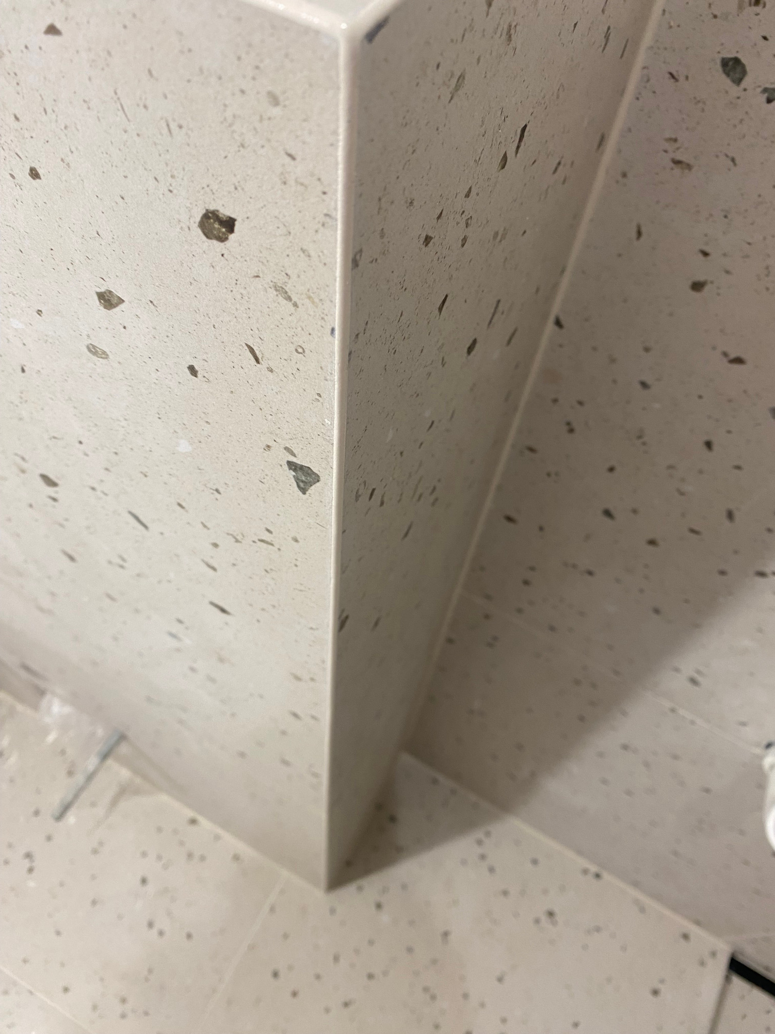 Narożnik obudowy wanny lub brodzika z jasnych płytek lastryko, widoczna fuga, minimalny fragment podłogi z tego samego materiału.