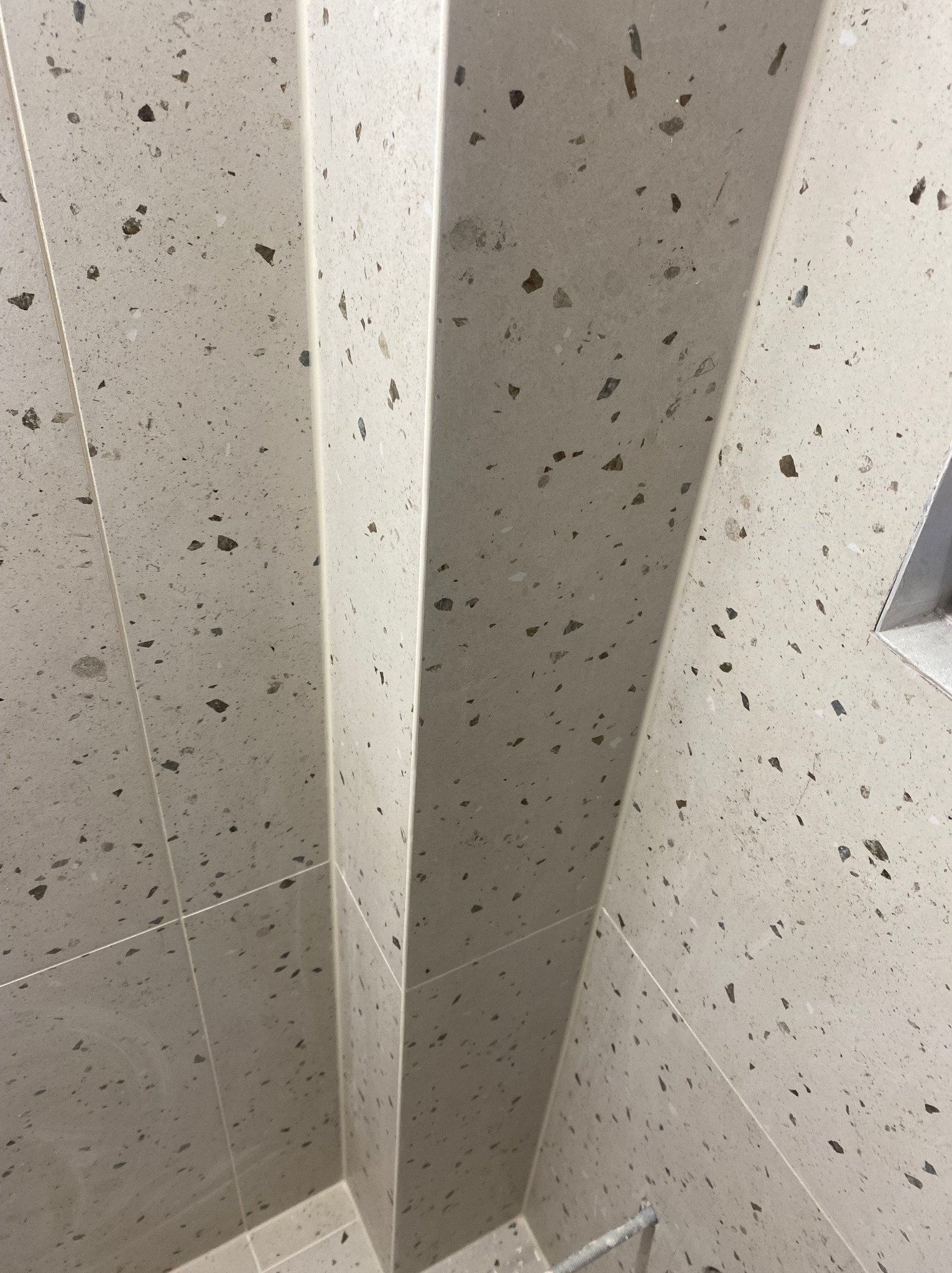 Beżowe, wielkoformatowe płytki imitujące lastryko, ułożone na ścianie i podłodze w narożniku kabiny prysznicowej, z widocznymi fugami.