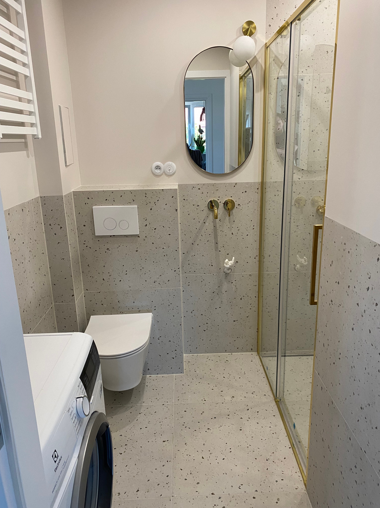 Nowoczesna łazienka z szarymi płytkami lastryko, złotymi akcentami w postaci ramy kabiny prysznicowej i baterii, owalnym lustrem z kulistym kinkietem, białą toaletą podwieszaną i pralką Electrolux.