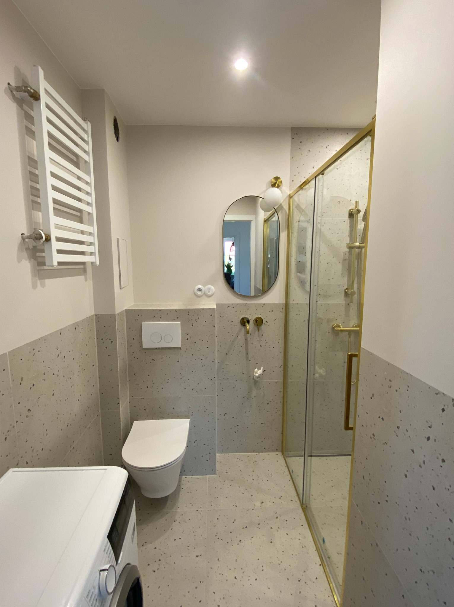 Nowoczesna łazienka z kabiną prysznicową ze złotymi ramami, owalnym lustrem z oświetleniem, umywalką, toaletą podwieszaną i pralką, wykończona płytkami imitującymi lastryko.