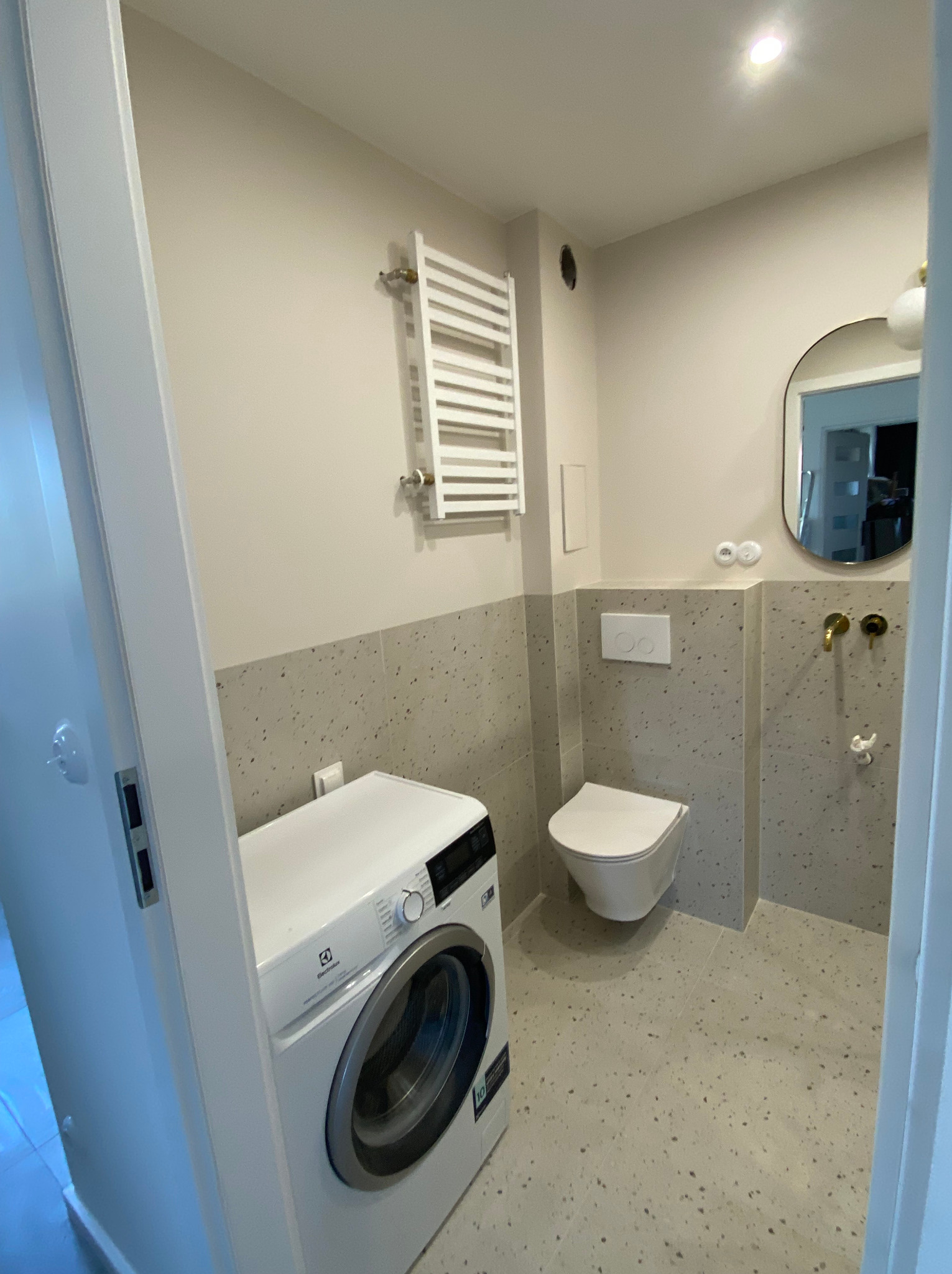 Nowoczesna łazienka w jasnych barwach z pralką Electrolux, toaletą podwieszaną i grzejnikiem drabinkowym, wykończona płytkami imitującymi lastryko.