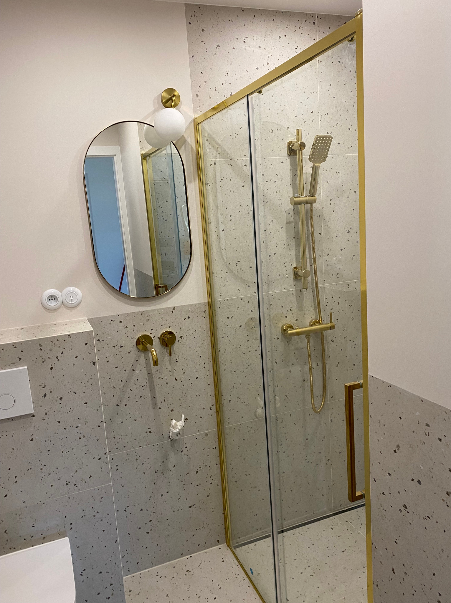 Nowoczesna łazienka z kabiną prysznicową w złotej ramie, owalne lustro z kulistym oświetleniem, bateria umywalkowa i płytki w stylu lastryko.