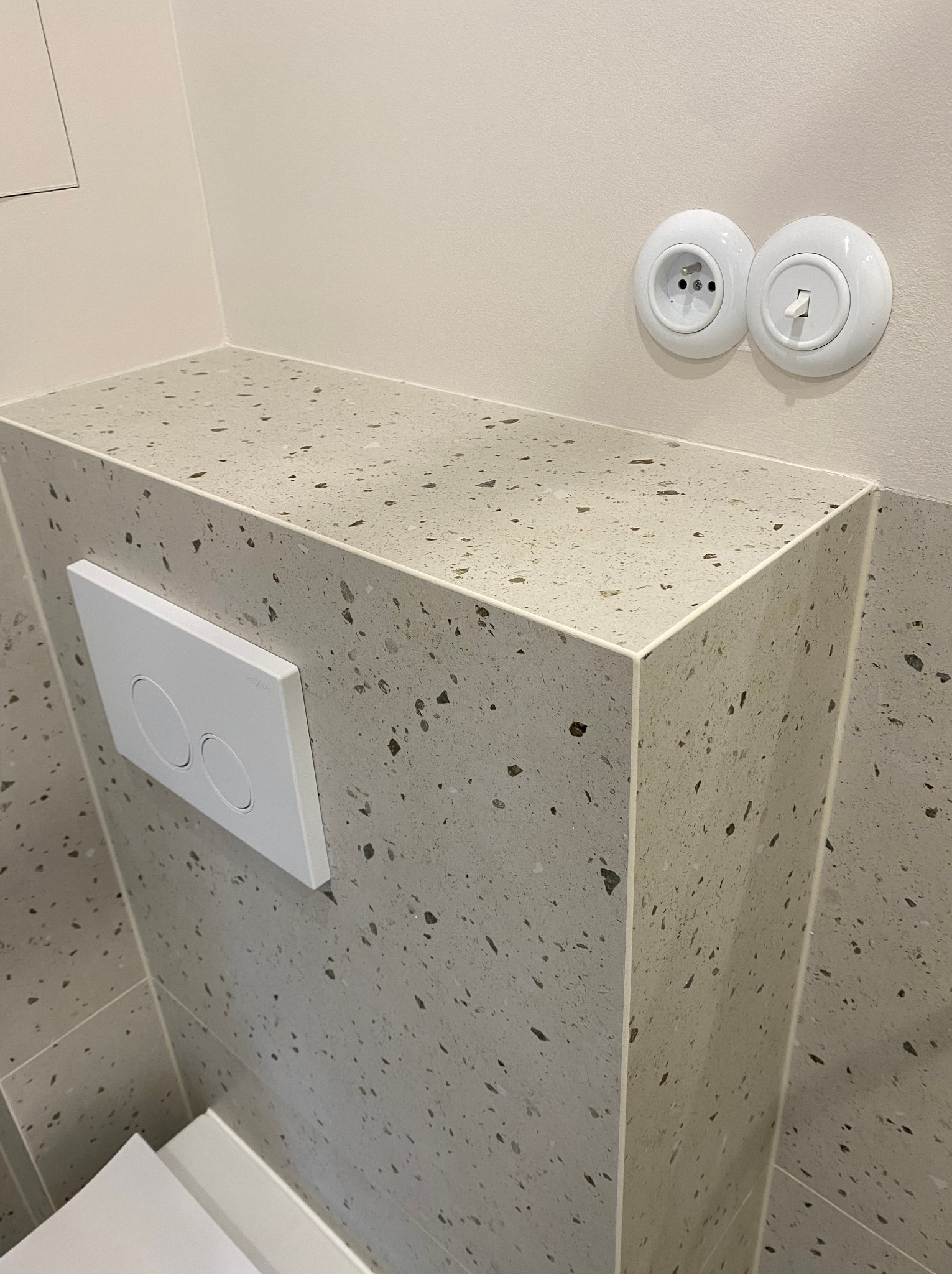 Fragment łazienki po remoncie: zabudowa stelaża WC wykończona płytkami lastryko, biały przycisk spłukujący Geberit, okrągłe gniazdko i włącznik retro na beżowej ścianie.