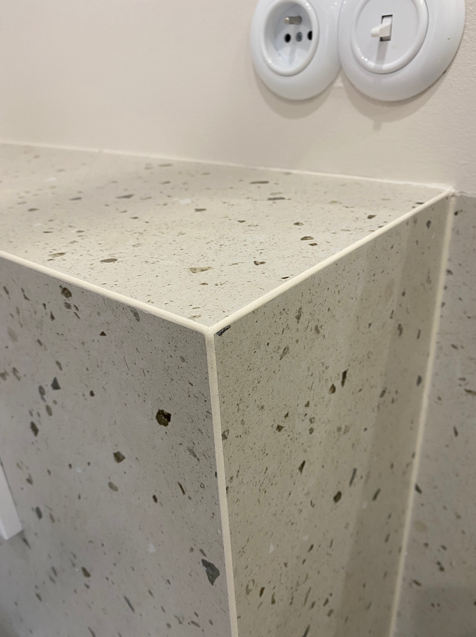 Fragment blatu łazienkowego z konglomeratu kwarcowego o nieregularnym wzorze, widoczne gniazdko i włącznik światła w stylu retro na ścianie.