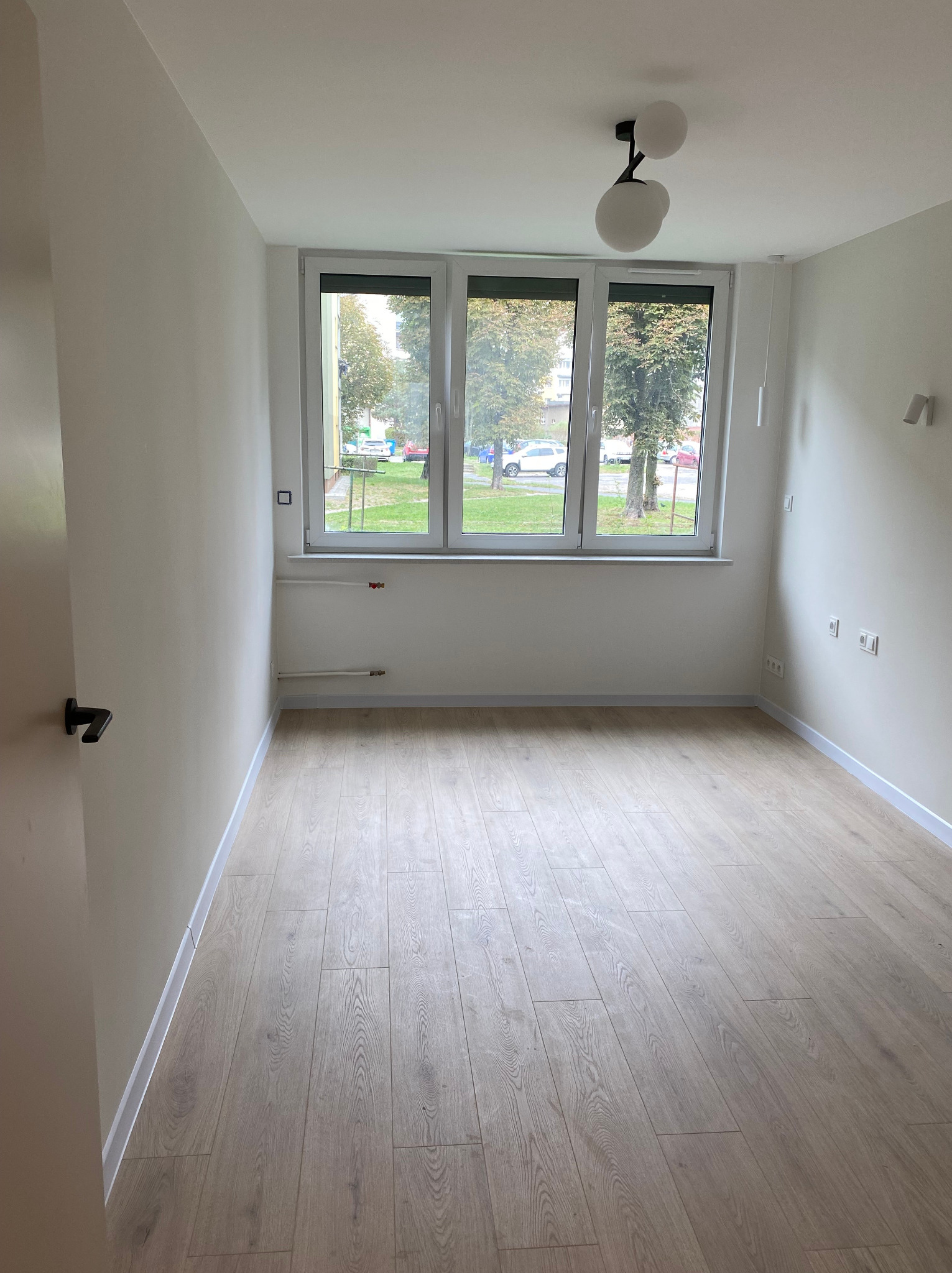 Pusty pokój z nową podłogą z paneli, świeżo pomalowanymi ścianami na biało i trzema oknami z widokiem na zieleń na zewnątrz.