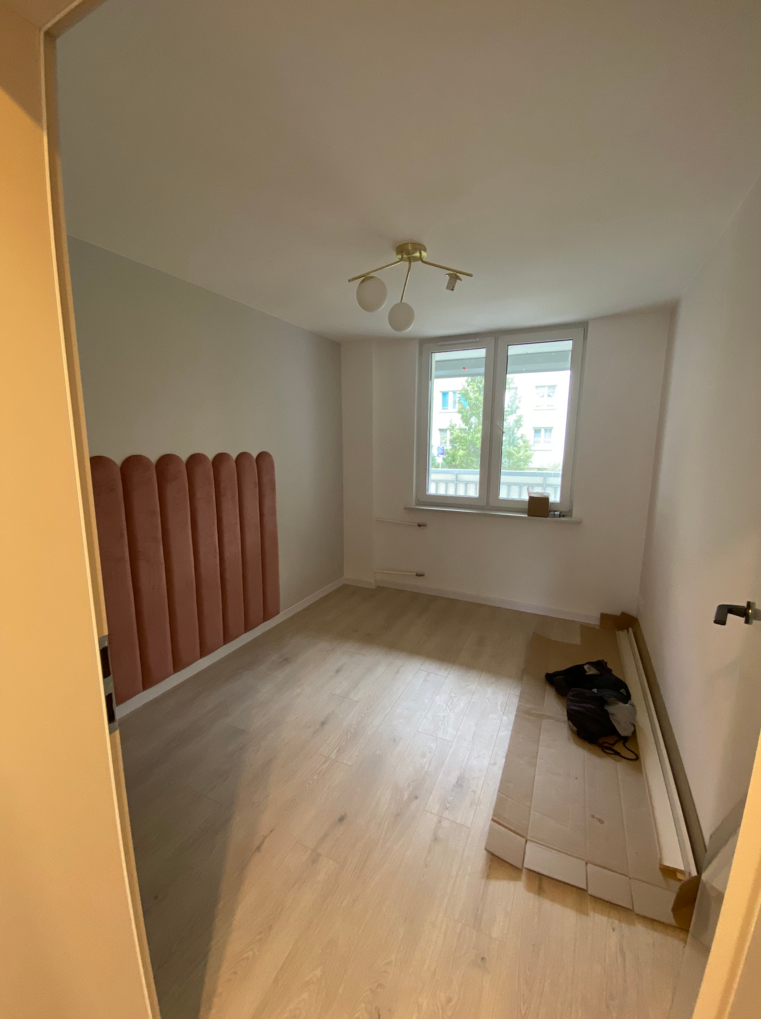 Pusty pokój z panelami ściennymi w kolorze pudrowego różu, drewnianą podłogą i białymi ścianami, widok zza uchylonych drzwi, na podłodze leżą profile przypodłogowe i torba.