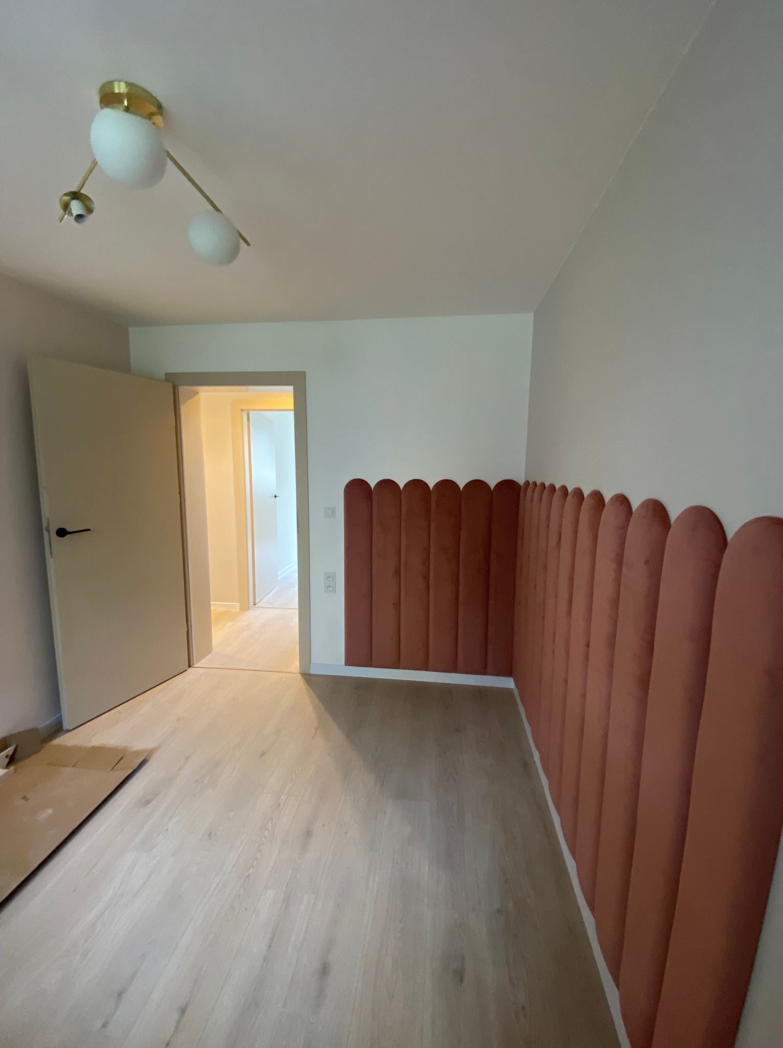 Pusty pokój z jasną podłogą i dekoracyjnymi, pionowymi panelami ściennymi w odcieniach terakoty, zakończonymi zaokrąglonymi brzegami, widoczne niedokończone podłogi i otwarte drzwi.