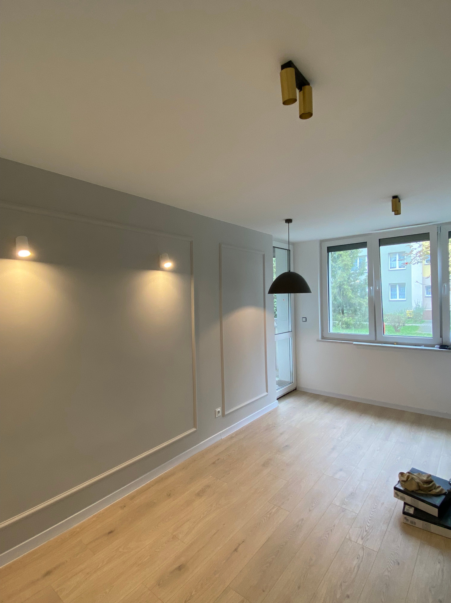 Wnętrze pokoju po remoncie: szare ściany z dekoracyjnymi listwami, jasna drewniana podłoga, nowoczesne oświetlenie sufitowe, widok na zieleń za oknem, kartony na podłodze.