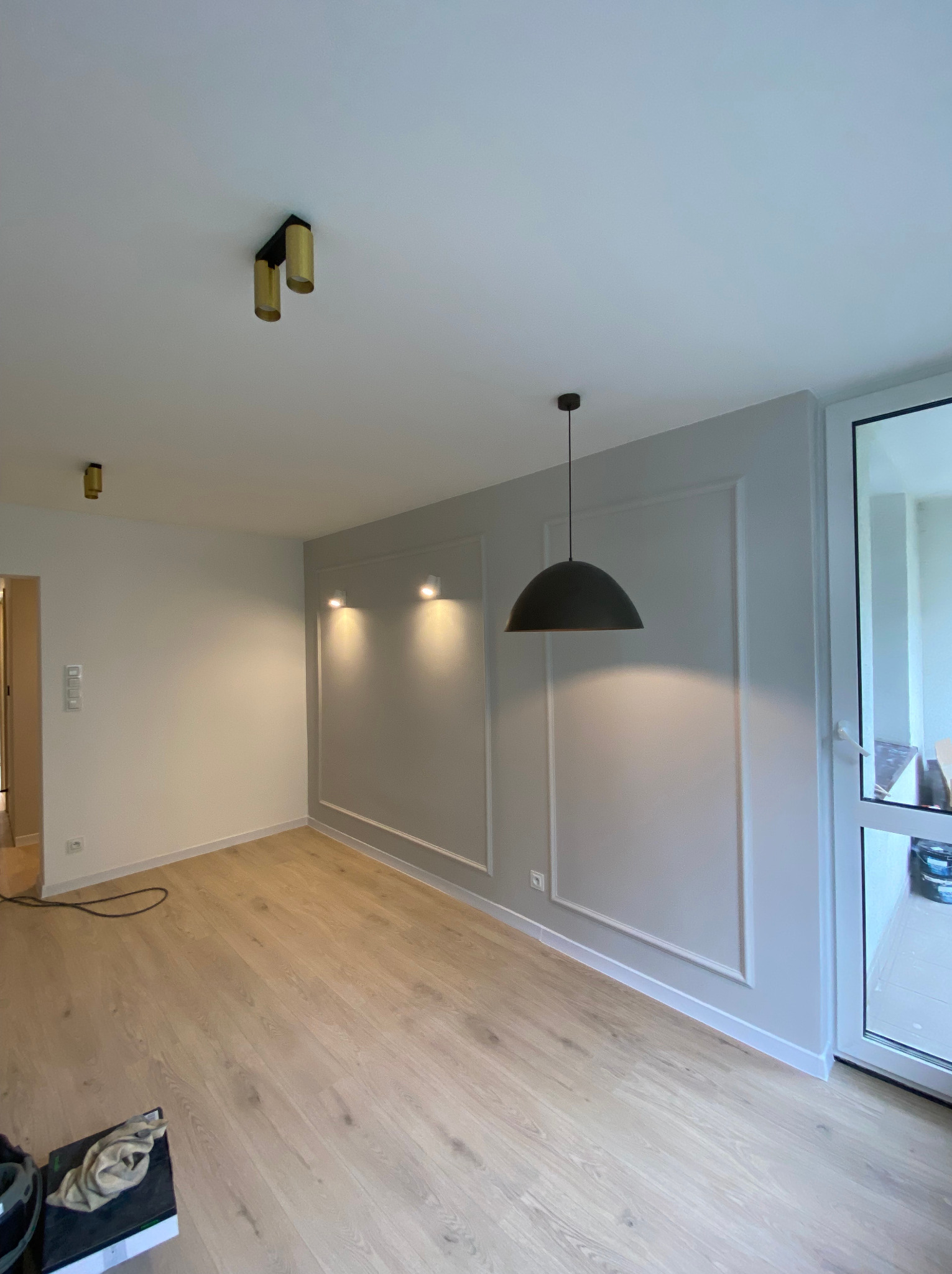 Pusty pokój po remoncie z panelami podłogowymi, szarą ścianą z dekoracyjnymi ramkami, nowoczesnym oświetleniem sufitowym i dużym oknem balkonowym.