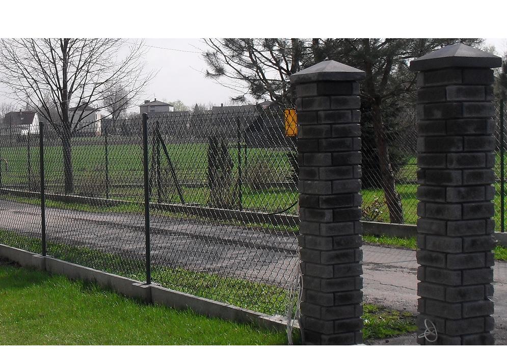 Siatka ogrodzeniowa z podmurówką betonową i słupkami z szarej cegły, widok na posesję z drzewami w tle.