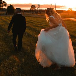 ArtBlur Julia Kipa - Para młoda idzie przez łąkę w promieniach zachodzącego słońca; panna młoda podnosi suknię, widoczny koronkowy tył sukni.