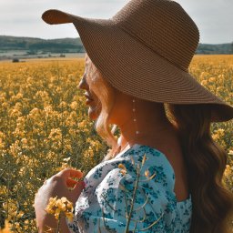 ArtBlur Julia Kipa - Kobieta w słomkowym kapeluszu i kwiecistej bluzce, stojąca bokiem na tle pola rzepaku, trzymająca delikatnie kwiatek w dłoni, z widocznymi perłowymi kolczykami.
