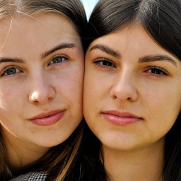 ArtBlur Julia Kipa - Portret dwóch młodych kobiet, z bliska, z delikatnym uśmiechem, włosy w odcieniach blondu i brązu, jedna w kraciastej marynarce, druga w czarnej bluzce z delikatnym złotym łańcuszkiem, na tle...