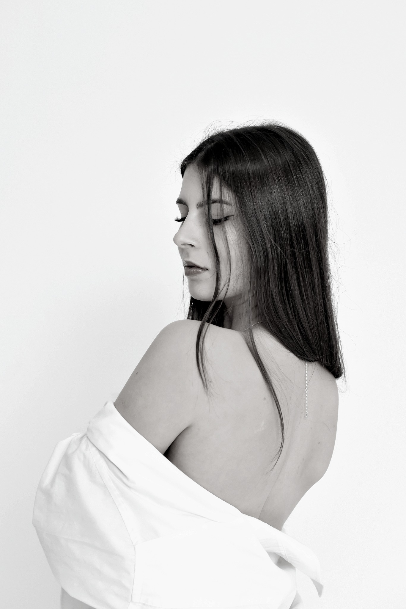 Portret w czerni i bieli kobiety z długimi, ciemnymi włosami, odwróconej tyłem, z opuszczoną białą koszulą eksponującą ramię i plecy, delikatny łańcuszek na szyi, spokojny wyraz twarzy, gładkie tło.