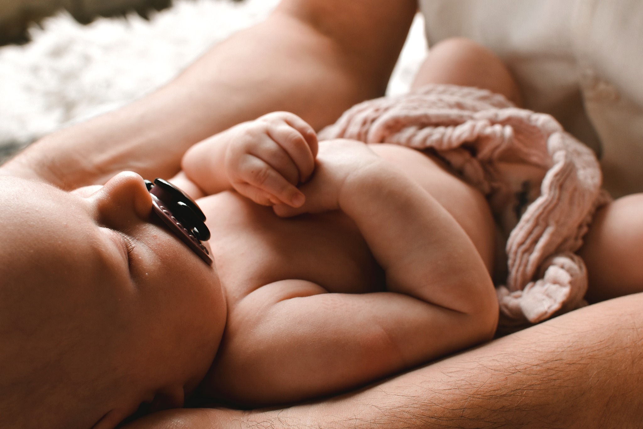 Śpiący noworodek z smoczkiem, owinięty w różowy kocyk, leży bezpiecznie w ramionach dorosłego.