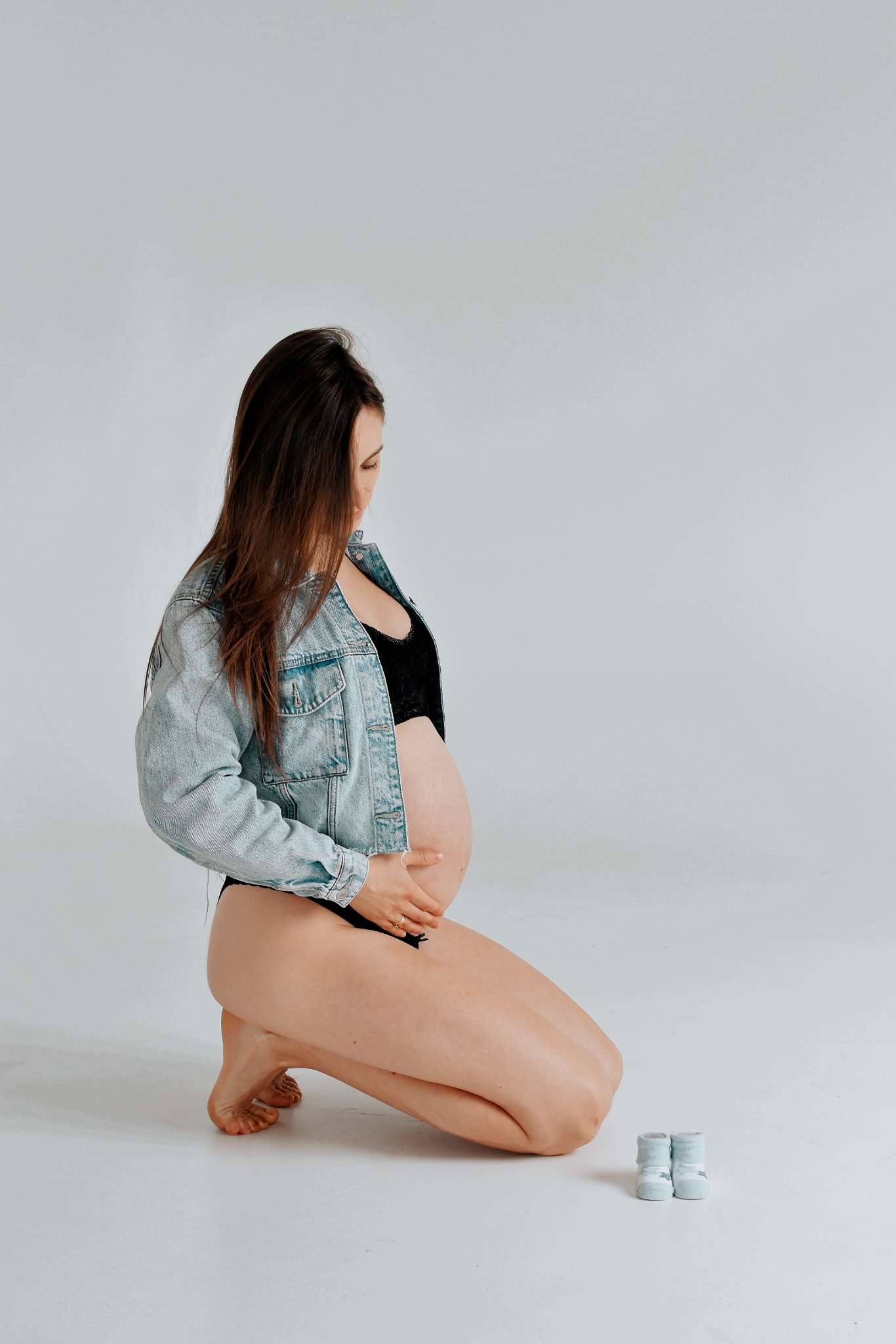 Kobieta w ciąży, w dżinsowej kurtce i czarnej bieliźnie, klęczy na białym tle, obok pary malutkich, niebieskich skarpetek.