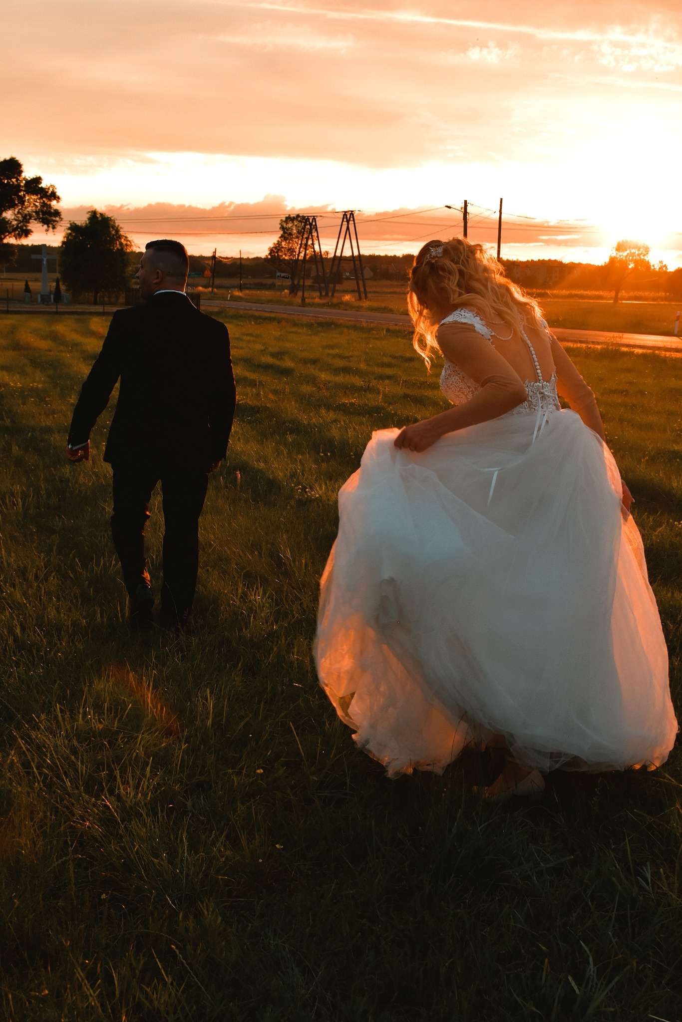 Para młoda idzie przez łąkę w promieniach zachodzącego słońca; panna młoda podnosi suknię, widoczny koronkowy tył sukni.