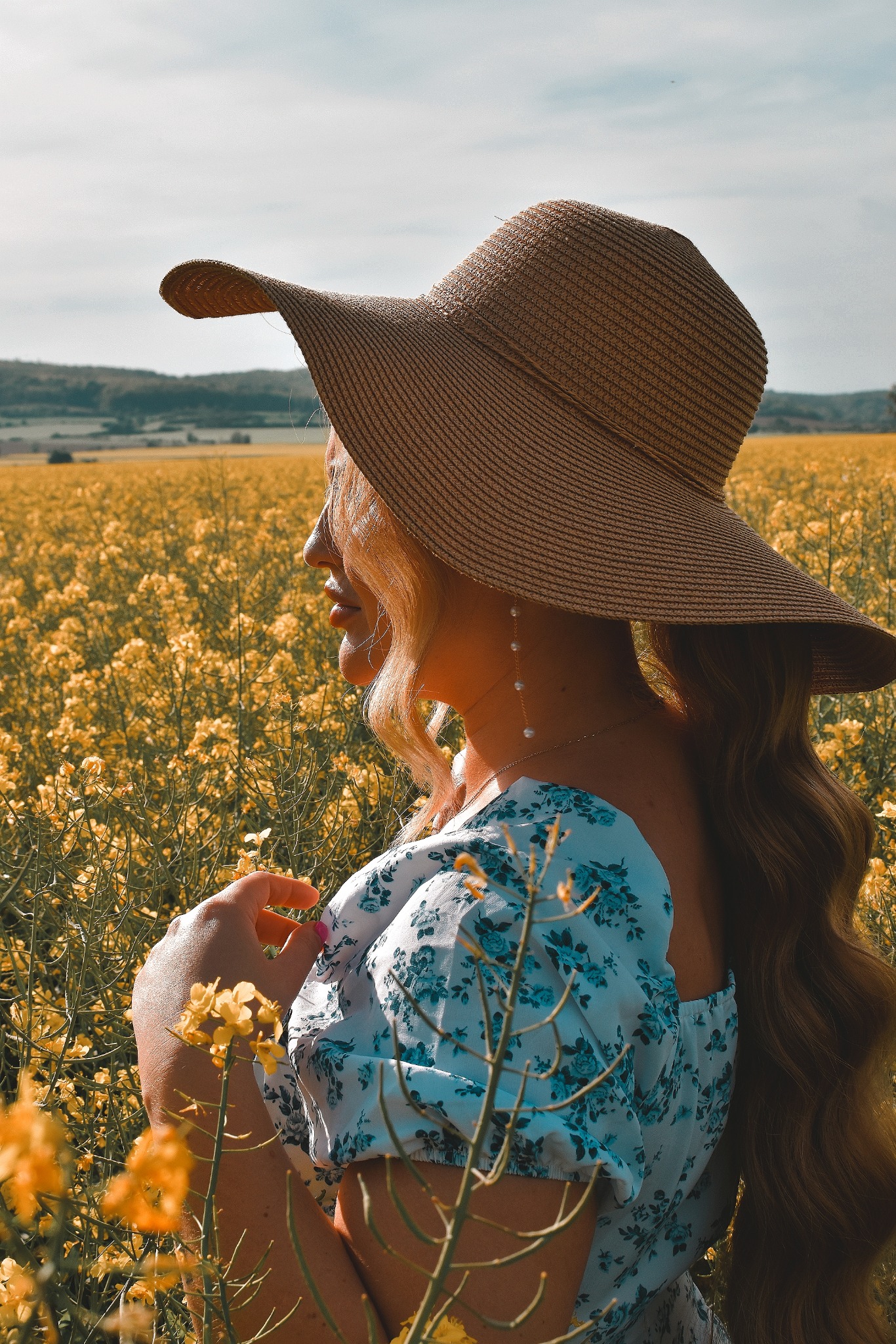 Kobieta w słomkowym kapeluszu i kwiecistej bluzce, stojąca bokiem na tle pola rzepaku, trzymająca delikatnie kwiatek w dłoni, z widocznymi perłowymi kolczykami.