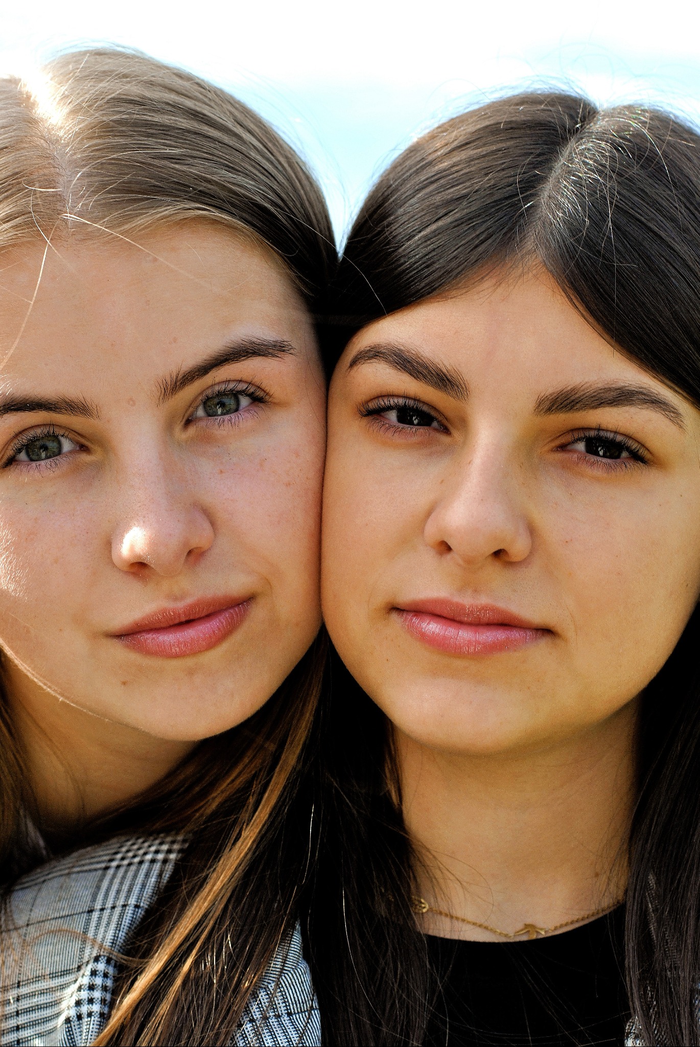 Portret dwóch młodych kobiet, z bliska, z delikatnym uśmiechem, włosy w odcieniach blondu i brązu, jedna w kraciastej marynarce, druga w czarnej bluzce z delikatnym złotym łańcuszkiem, na tle...