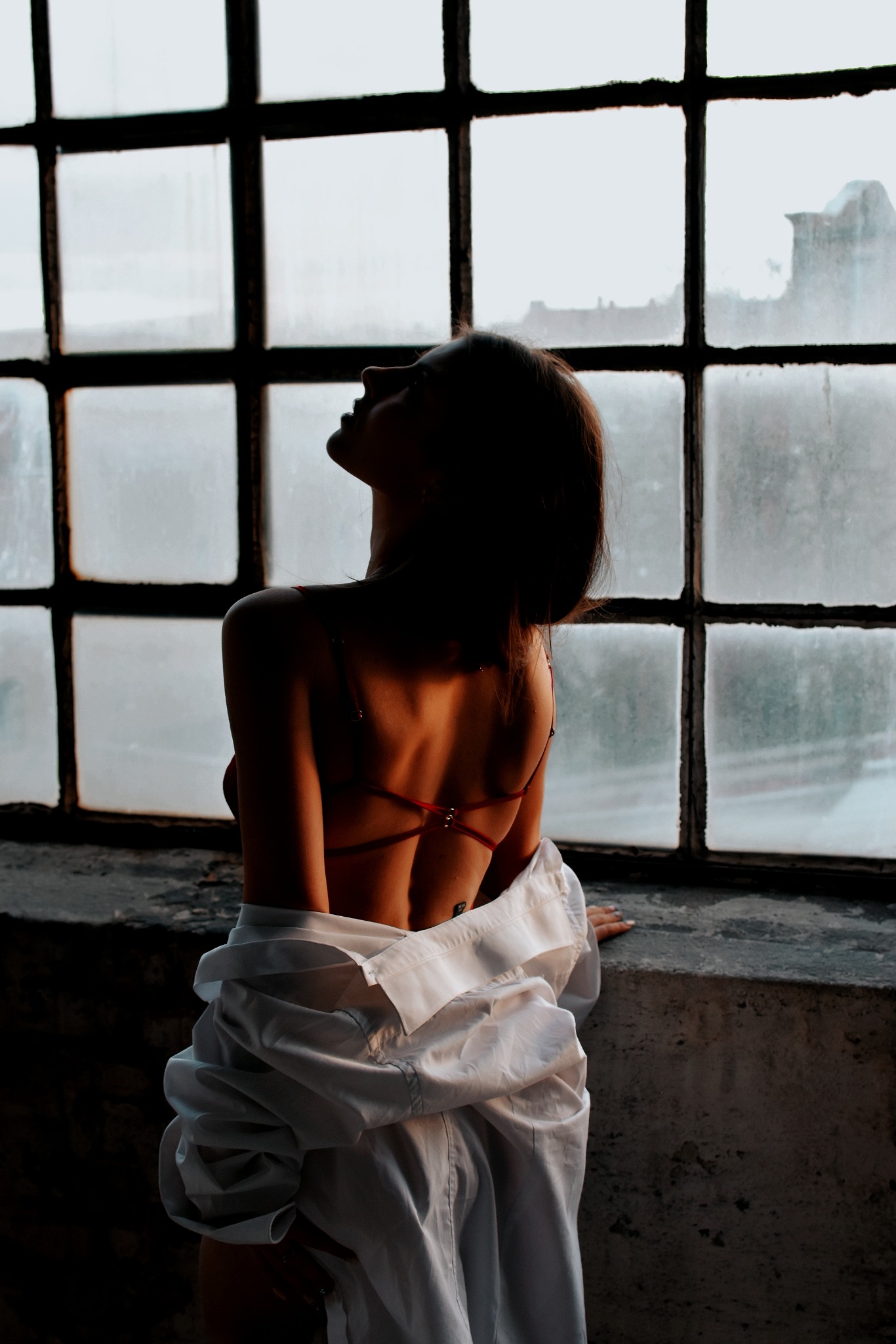 Kobieta w bieliźnie i rozpiętej białej koszuli, odwrócona tyłem, spogląda w górę na okno z grubymi, ciemnymi ramami i rozmytym widokiem za szybą. Na plecach widoczny mały tatuaż.