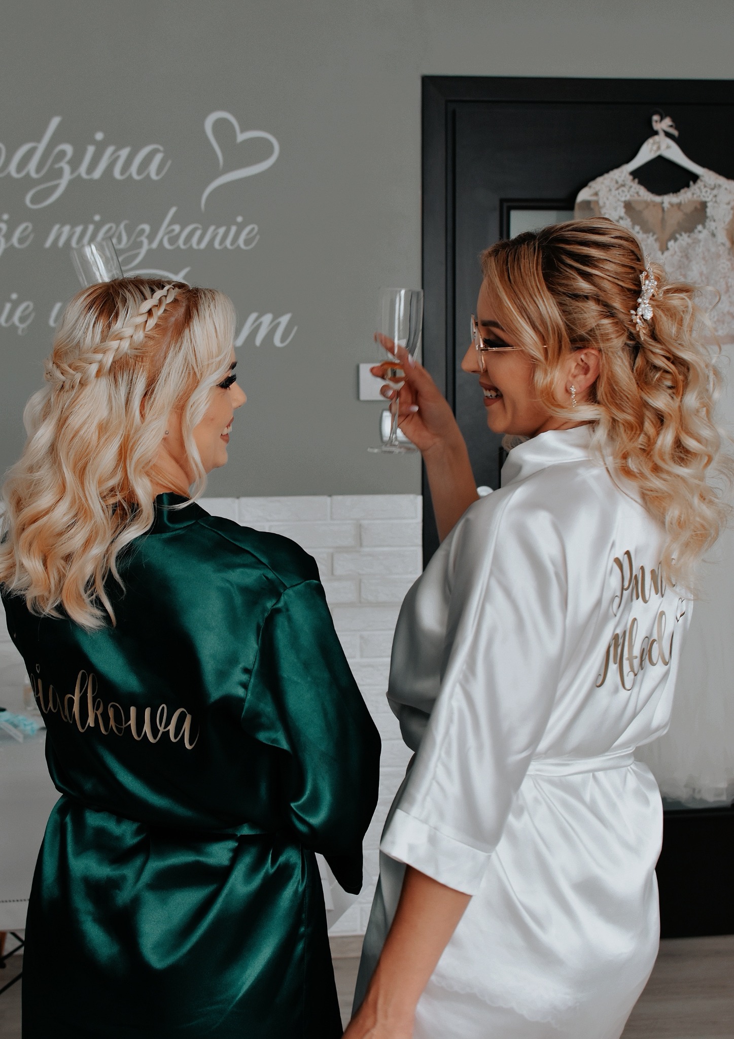 Dwie kobiety w szlafrokach, panna młoda w białym z napisem 'Panna Młoda' na plecach, druhny w szmaragdowym z napisem 'Świadkowa', wznoszą toast kieliszkami, w tle suknia ślubna na wieszaku i napis...