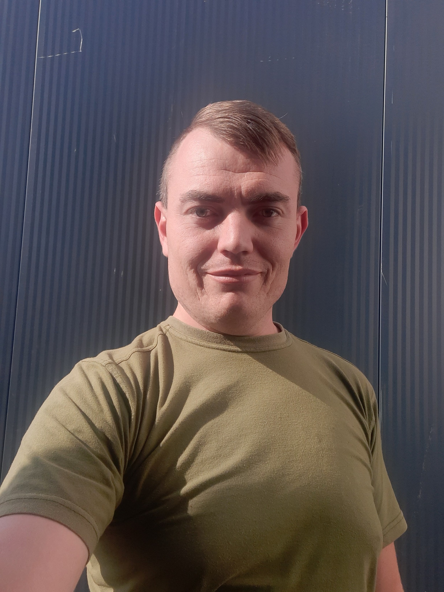 Uśmiechnięty mężczyzna w zielonej koszulce na tle pionowo żłobkowanej, ciemnoniebieskiej ściany z lekkim zarysowaniem.