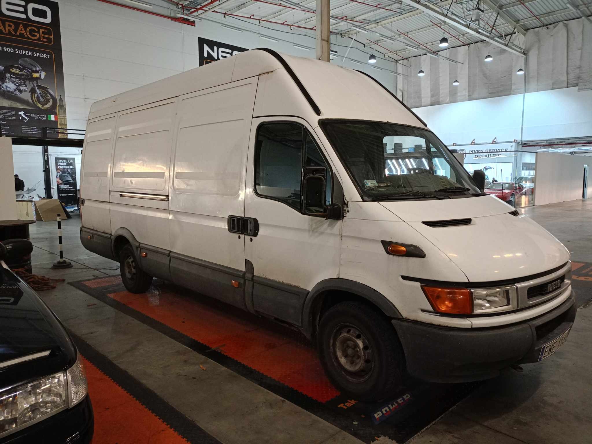 Biały, używany furgon marki Iveco zaparkowany w garażu w Warszawie, widoczny z przodu i boku, z widocznymi śladami użytkowania i zabrudzeniami.
